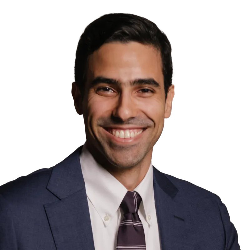 A professional headshot of a man wearing a dark blue suit, white shirt, and striped tie. He has short dark hair, a wide smile, and is looking directly at the camera against a transparent background.